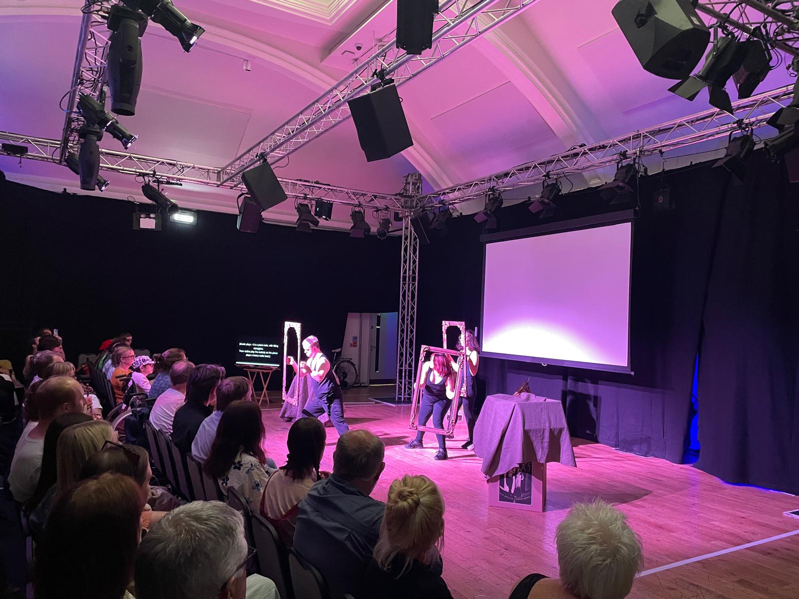 Performers act on stage using large frame props while a full audience watches from closely packed seats.