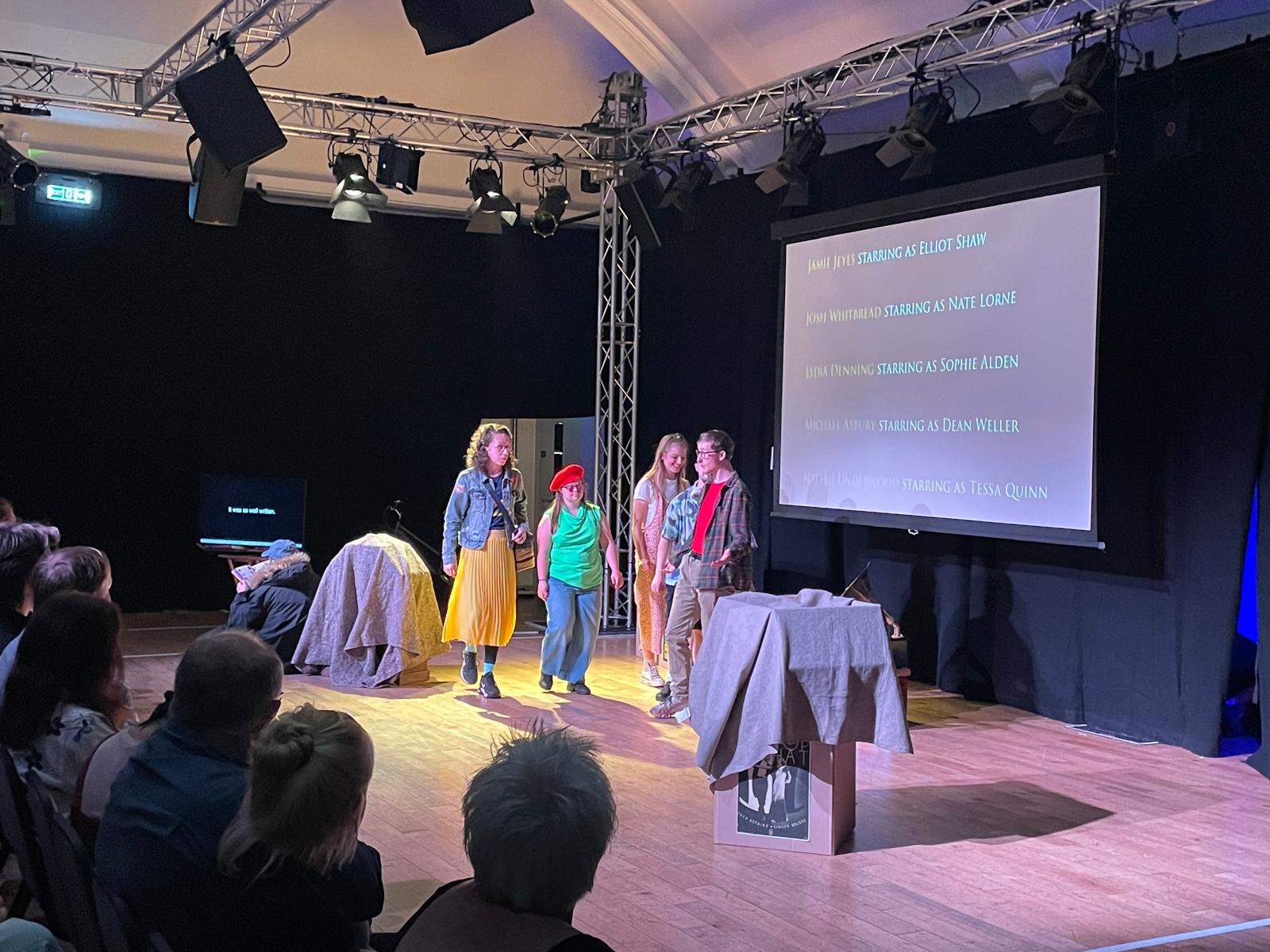Members of the Reel to Real cast perform on stage during the final show, with audience members visible in the foreground and cast credits projected on the screen behind them.