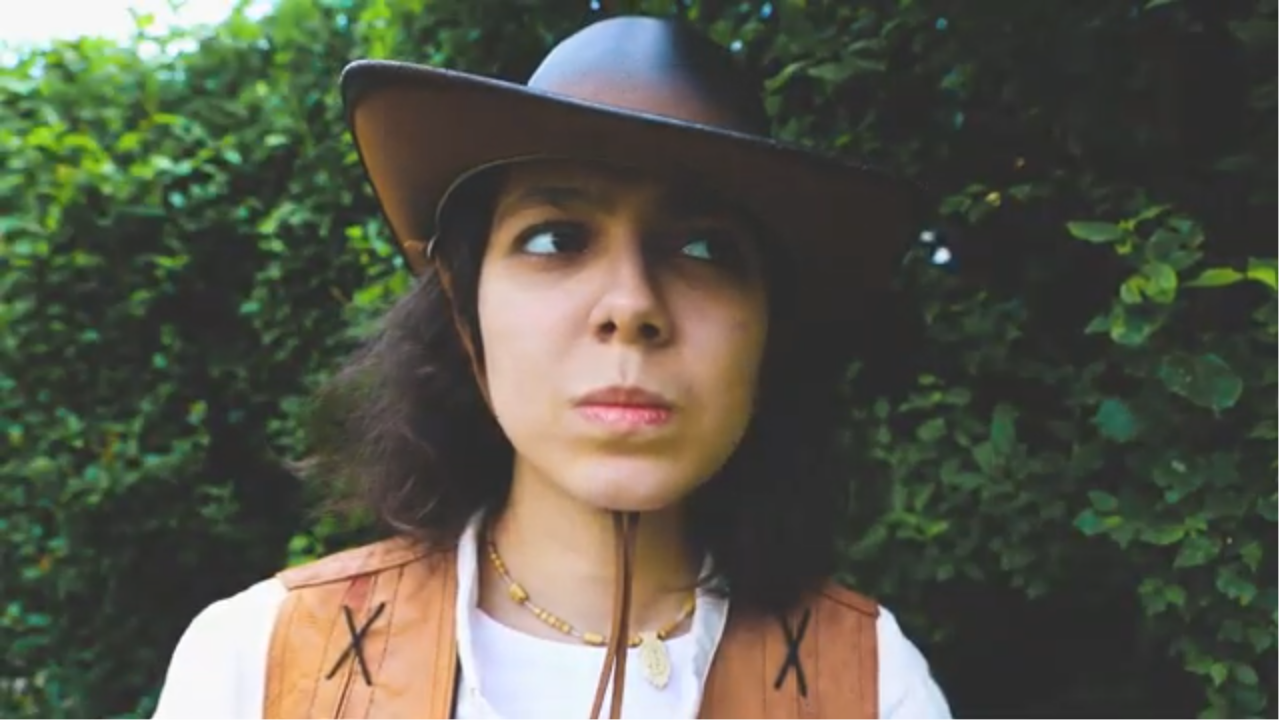A close up on Chrissie, wearing a cowboy hat and waistcoat, looking intense.