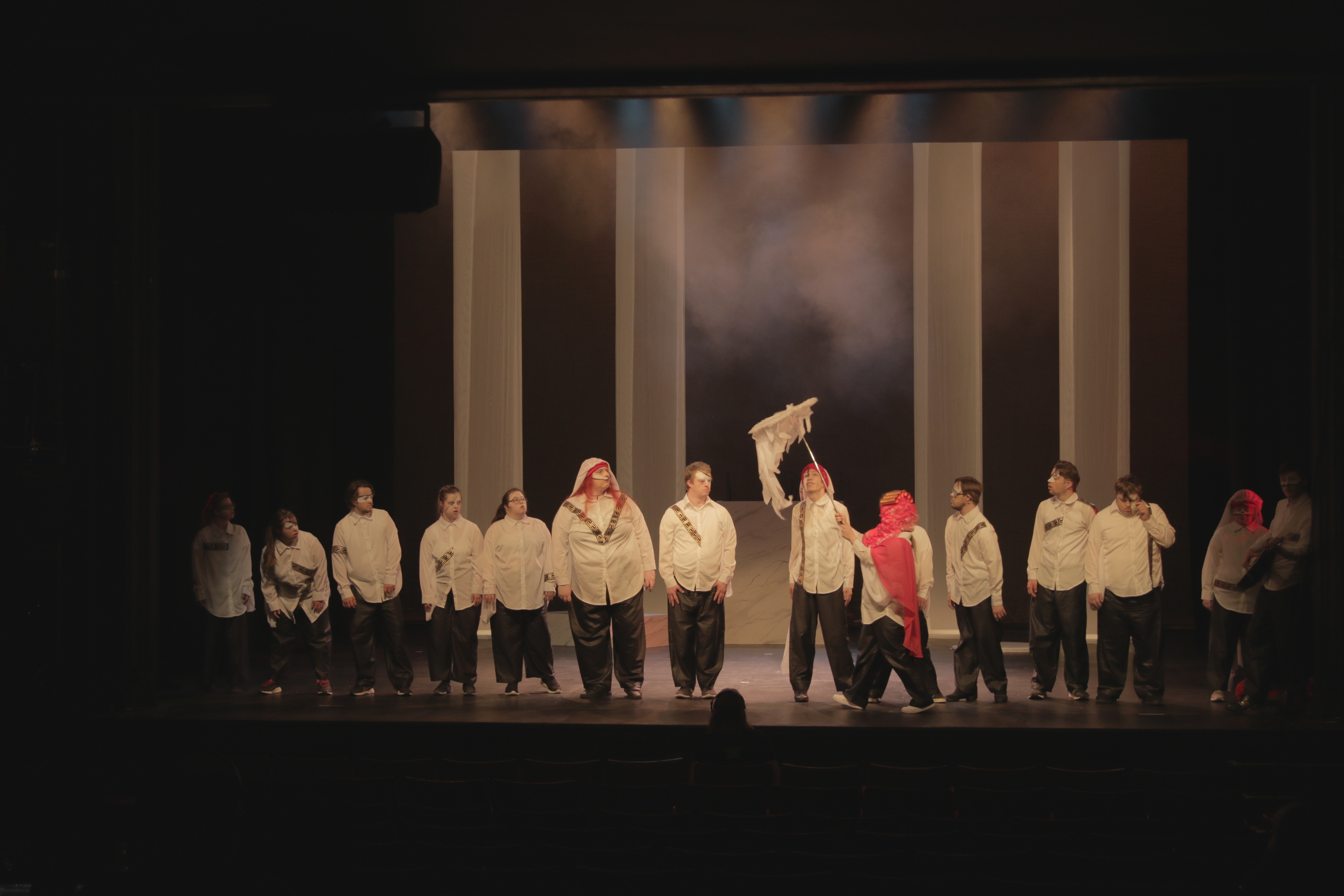Wide stage photo showing a group of performers in white shirts and dark trousers lined up across the stage during a theatrical performance, with one performer in red moving through the centre and another holding a white prop beneath a misty, atmospheric backdrop.