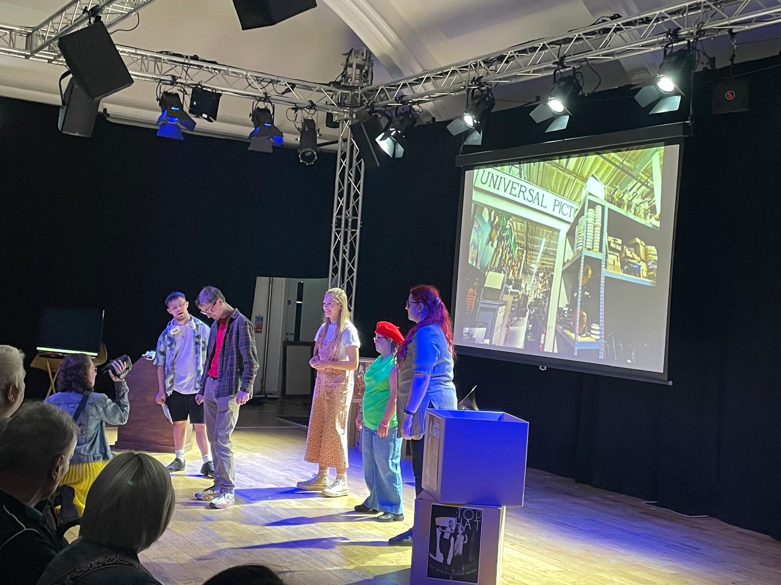 Members of the Reel to Real cast perform on stage under theatre lighting, with a projected image behind them and audience members watching from the foreground.