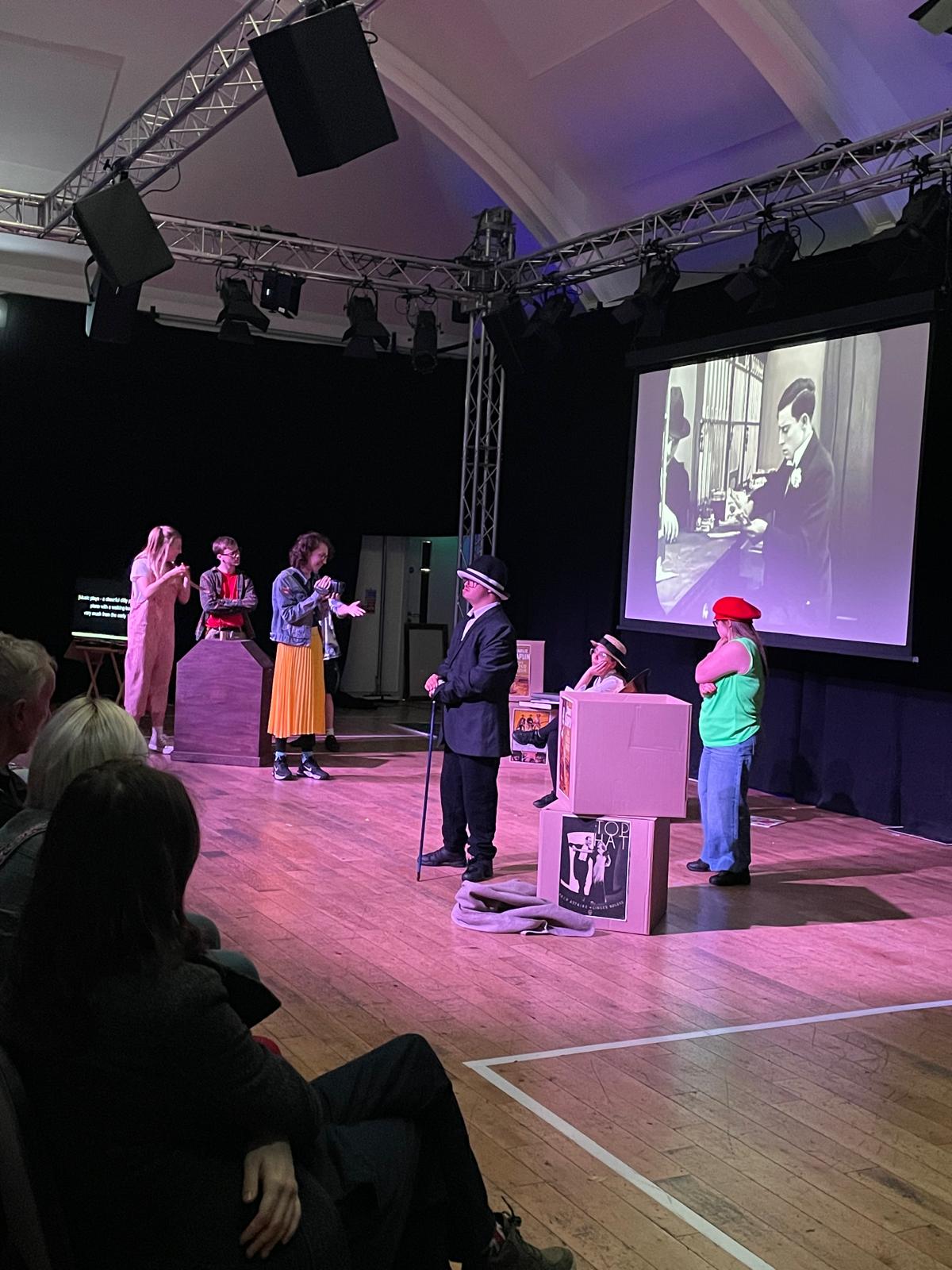 Several performers act out a scene on stage while a black-and-white silent film image is projected onto the large screen behind them.