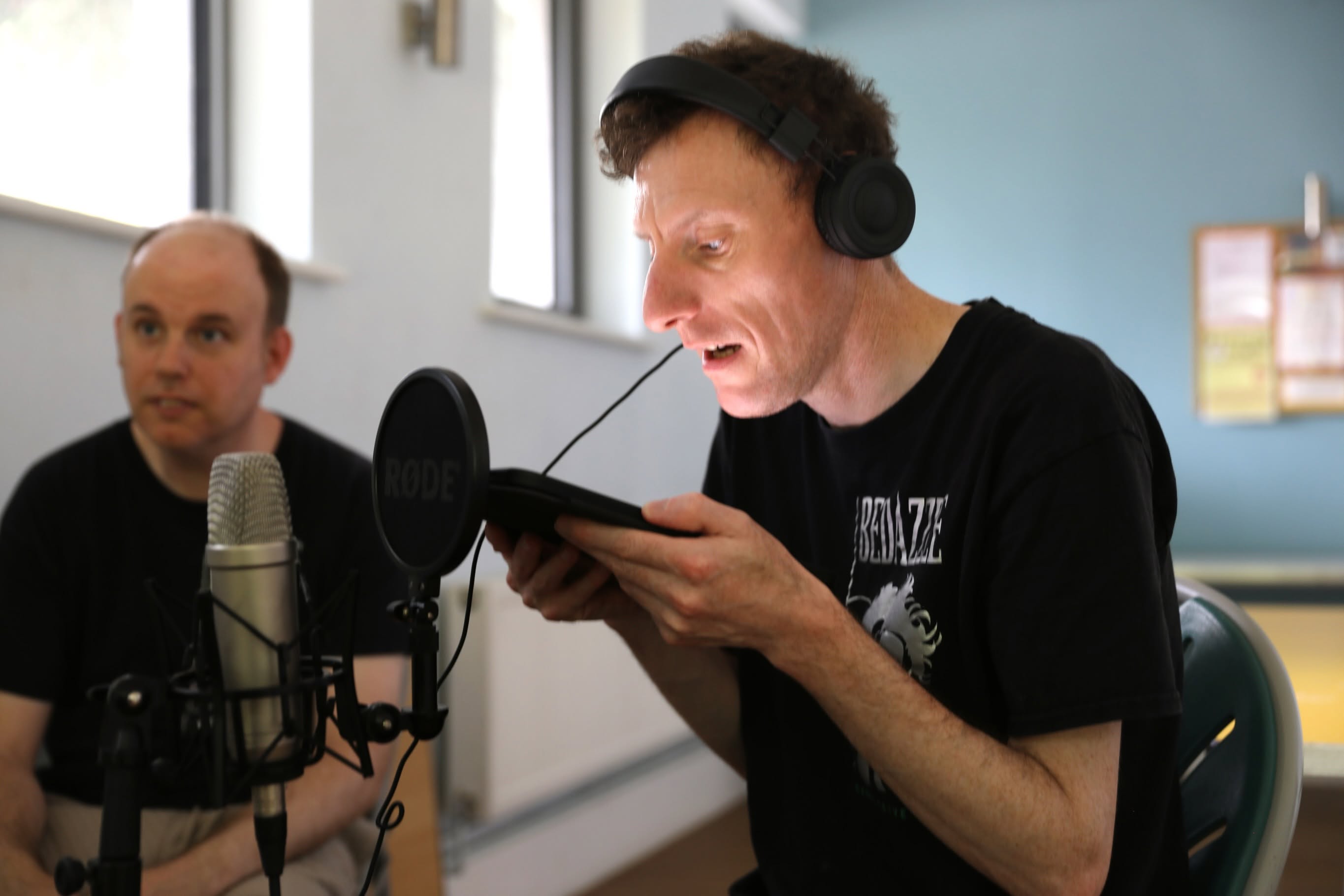 A Bedazzle performer wearing headphones speaks into a microphone while reading from a tablet, with another performer seated beside him.